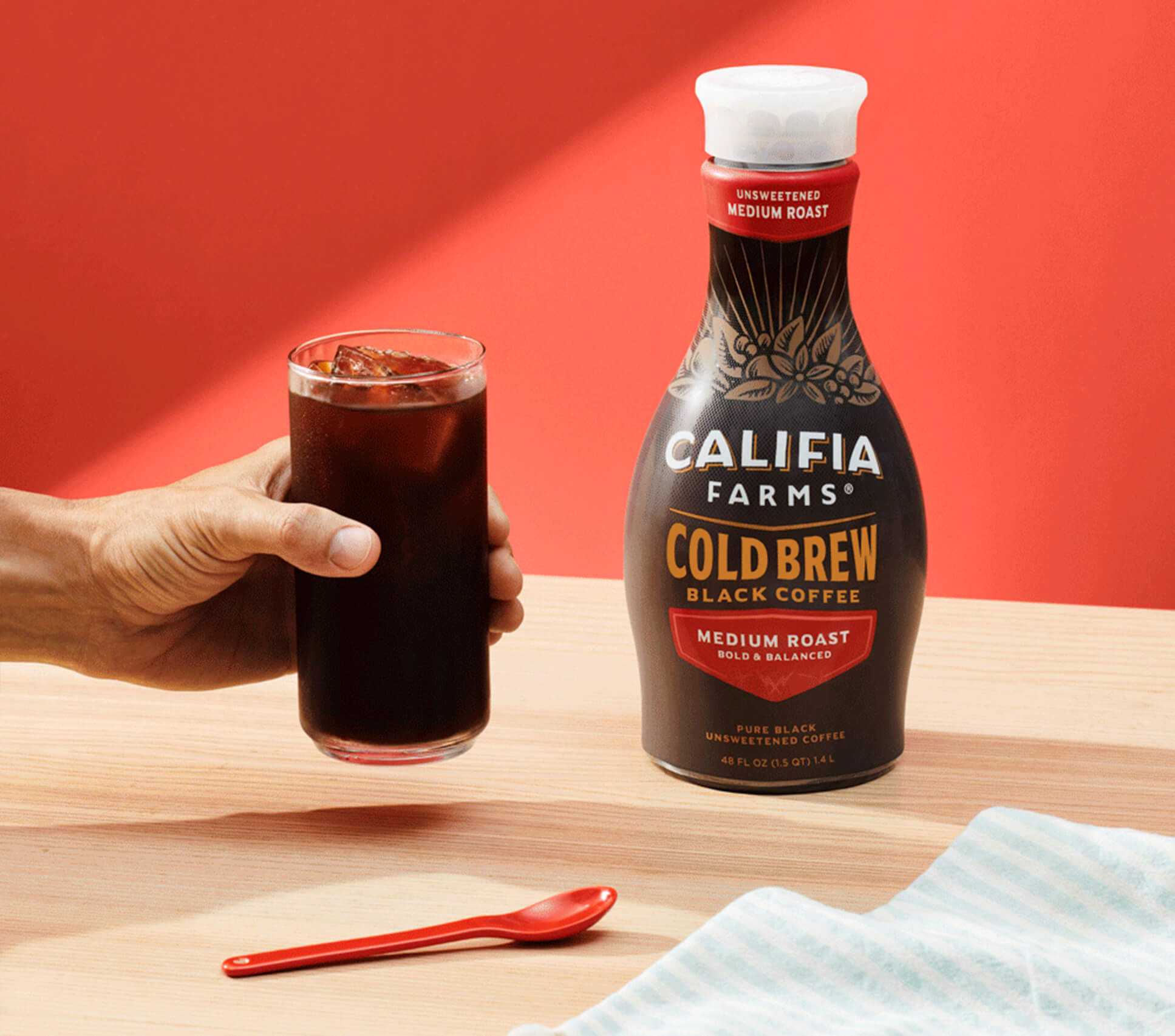 a close-up of a hand holding a glass of iced coffee with Califia Cold Brew in the background